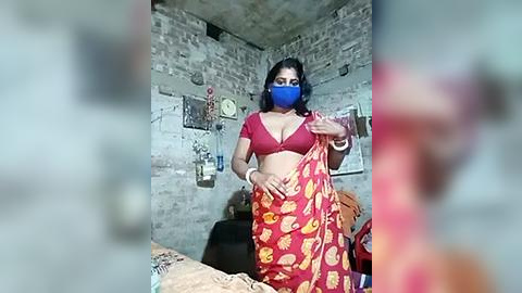 Media: A video of a woman with medium skin tone, wearing a red bra and red sari with yellow patterns, blue mask, and bracelets, standing in a rustic room with a brick wall.