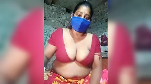 Media: Video of a plus-sized woman with medium skin tone, wearing a red top, blue mask, and colorful saree, indoors with a rustic background.