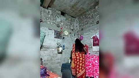 Media: Video of a woman in a red sari with white polka dots, standing in a dimly lit, rustic room with exposed brick walls, hanging clothes, and a clock on the wall.