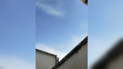 Media: A video capturing a narrow, blue sky with wispy clouds above a beige building with a tiled roof and a dark metal railing in the foreground. The image is slightly out of focus, creating a dreamy, slightly blurred effect.