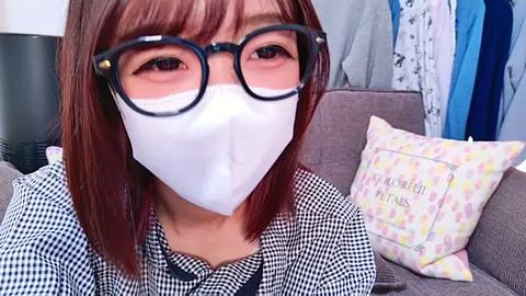 Media: A video of an East Asian woman with straight, shoulder-length brown hair, wearing black-rimmed glasses, a white face mask, and a black-and-white checkered shirt, sitting on a gray couch with floral pillows, in a brightly lit room.