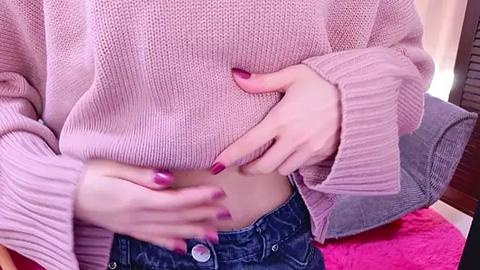 Media: A video of a person wearing a pink knit sweater, exposing their midriff, with hands resting on their stomach. The person's nails are painted pink. The background features a wooden floor and a pink rug.