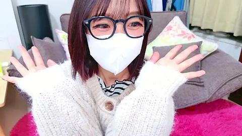 Media: Video of an Asian woman with shoulder-length brown hair, wearing glasses, a white mask, and a light knit sweater, seated on a couch with cushions and a pink blanket, indoors.