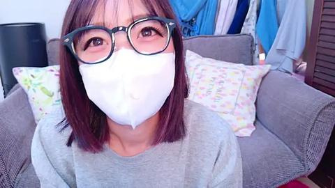 Media: A video of an Asian woman with shoulder-length brown hair, wearing glasses and a white face mask, sitting on a gray couch with colorful pillows. The background shows a cluttered room with blue fabric and wooden furniture.