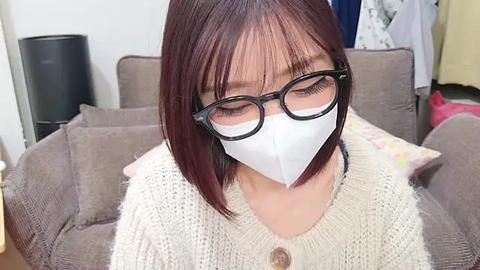 Media: Video of an Asian woman with short, straight auburn hair, wearing black-rimmed glasses, a white face mask, and a cream-colored, knitted sweater. She sits on a gray couch in a living room with a black cylindrical lamp and a white wall.