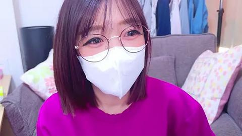 Media: A video of an Asian woman with shoulder-length brown hair, wearing a white face mask, round glasses, and a pink shirt. She is indoors, in front of a gray couch with colorful pillows, and a blue garment hangs on a wall.