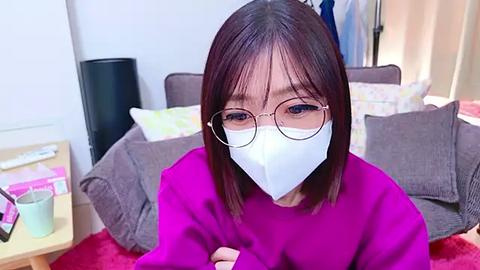 Media: A video of an Asian woman with straight, shoulder-length black hair, wearing glasses and a white surgical mask, sitting on a red couch in a living room with a grey chair and a pink blanket.