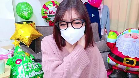 Media: A video of an East Asian woman with shoulder-length hair, wearing black-framed glasses and a white face mask, seated in a living room adorned with festive decorations, including a red Santa hat and a green balloon.