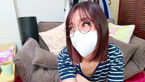 Media: Video of a young woman with shoulder-length brown hair, wearing glasses and a white mask, sitting on a beige couch in a cozy living room.