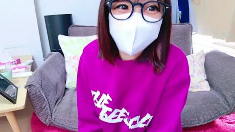 Media: Video of a woman with short brown hair, wearing black-framed glasses, a white mask, and a vibrant pink sweatshirt with Japanese kanji. She sits on a grey couch in a cozy living room with a pink rug.