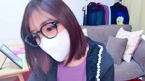 Media: Video of an Asian woman with shoulder-length dark hair, wearing black-rimmed glasses, a purple top, and a white mask, sitting on a gray sofa with a pink cushion. Background shows a cluttered room with a table, bags, and a TV.