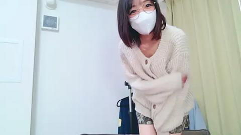 Media: A young East Asian woman with shoulder-length dark hair, glasses, and a white mask, wearing a cream cardigan and patterned shorts, leans forward in a room with white walls, a door, and a curtain.