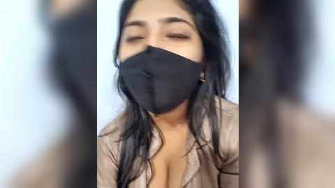 Media: Video of a woman with long, straight black hair, wearing a black face mask and a light brown jacket, eyes closed, blurred background.