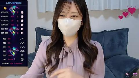 Media: A video of an Asian woman with long dark hair, wearing a white mask and light pink blouse, sitting on a blue couch. Background shows a TV screen with game statistics.