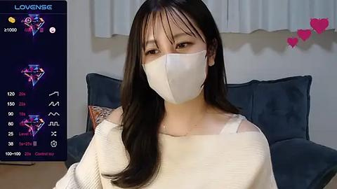 Media: Video of an Asian woman with long black hair, wearing a white face mask, beige sweater, and sitting on a dark blue sofa, with a \"LoveSense\" app screen showing her stats on the left.