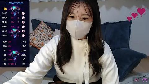 Media: A video of an East Asian woman with long black hair, wearing a white mask and white top, seated in a room with blue cushions and floral pillows.