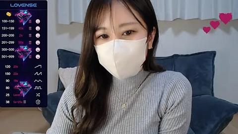 Media: Video of an Asian woman with long dark hair, wearing a white mask and a gray ribbed sweater, sitting indoors, with a video game screen overlay showing health and damage statistics.