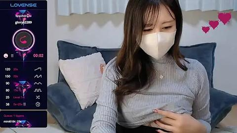 Media: Video of an East Asian woman with long black hair, wearing a white mask and a grey ribbed turtleneck, sitting on a grey couch. Background shows a digital screen with health statistics and a heart icon.