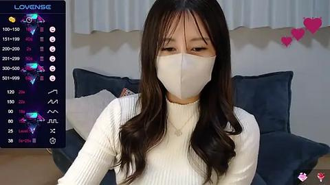 Media: Video of an East Asian woman with long, wavy brown hair, wearing a white face mask and a ribbed white turtleneck, seated in a living room with a blue sofa and pink heart decorations on the wall.