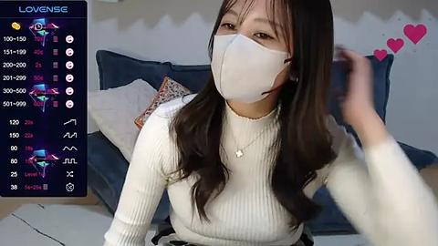 Media: A video of a young Asian woman with long black hair and fair skin, wearing a white face mask, white ribbed sweater, and a necklace. She sits on a couch, in a cozy room with a TV screen displaying virtual avatars.