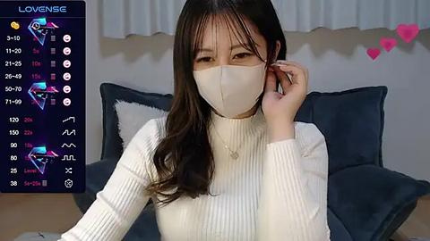 Media: Video of an East Asian woman with long black hair, wearing a white face mask, white ribbed sweater, and sitting on a bed with blue pillows. Background shows a digital screen with a \"Lovense\" app interface.