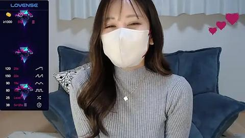 Media: Video of an Asian woman with long dark hair, wearing a white face mask and a grey ribbed top, seated in a blue-cushioned room with white curtains.