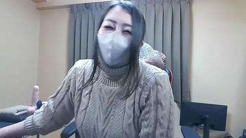 Media: Video of an Asian woman with long black hair wearing a beige knit sweater, face mask, and a thumbs-up gesture. Background includes a beige curtain and a computer monitor.