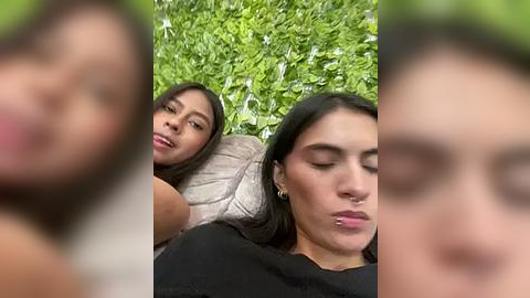 Media: Video of two young women lying on lush green grass, one with long brown hair, the other with dark hair. The background features blurred faces of other people.