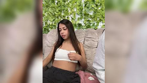 Media: Video of a young woman with long dark hair, wearing a white crop top and black pants, sitting on a beige couch with lush green vines in the background.