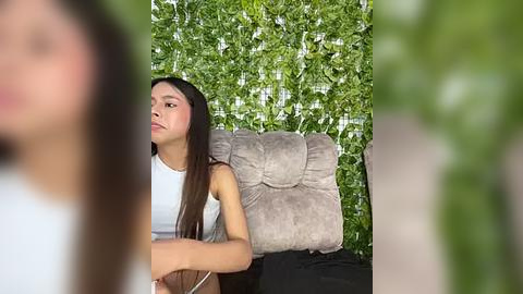 Media: Video of a young Asian woman with long, straight black hair, wearing a white tank top, sitting on a grey cushion against a green leafy backdrop.