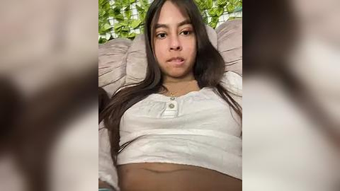 Media: Video of a young Latina woman with long dark hair, light skin tone, wearing a white crop top, lying on a cushioned surface with blurred background foliage.