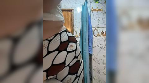 Media: Video of a woman in a bathroom, wearing a white and black patterned shirt. The background features a tiled wall, a wooden door, and a blue shower curtain.