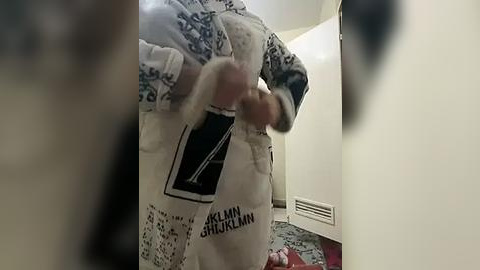 Media: Video of a person wearing a white apron with \"Geraldine J\u00fcrgens\" text and floral patterns, inside a dimly lit kitchen with beige walls, a patterned rug, and a white cabinet in the background.