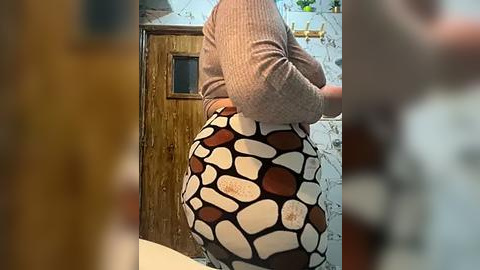 Media: Video of a person with a round, curvy body, wearing a beige sweater and a patterned skirt with stone-like shapes in white, brown, and black. The background features a wooden door and a tiled wall in a bathroom.