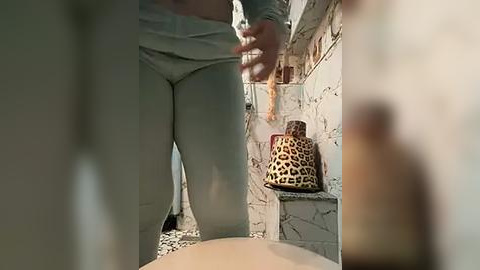 Media: Video of a person in a bathroom with light skin, wearing white pants and a leopard-print handbag, holding a phone. Marble tiles, a toilet, and a towel are visible.