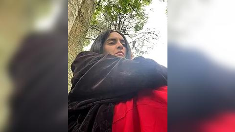 Media: A video of a young woman with long, dark hair, wearing a black velvet jacket over a red shirt, standing against a tree in a forest. The image is slightly blurred, emphasizing the natural surroundings.