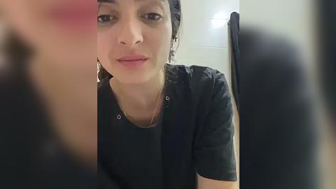 Media: A video of a young woman with olive skin, dark hair, and dark eyes, wearing a black shirt, looking into a mirror. The background shows a white tiled wall and a towel hanging on a rack.