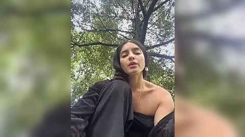 Media: Video of a young woman with medium-length black hair, wearing a black off-shoulder top, standing in a lush, green forest. Her expression is contemplative. The background features dense tree foliage.