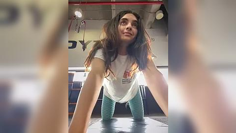 Media: A video of a young woman with long, wavy brown hair, wearing a white T-shirt and light blue leggings, performing a push-up on a gym mat. The background shows a dimly lit gym with red pipes and equipment.