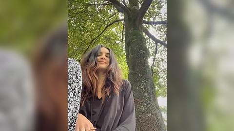 Media: Video of a woman with long, wavy brown hair, wearing a black robe, standing outdoors near a tree with green leaves, blurred background.