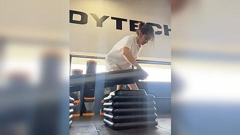 Media: Video of a woman with a bun wearing a white shirt squatting on a stack of weights in a gym. Background shows gym equipment and a \"DYTECH\" sign.