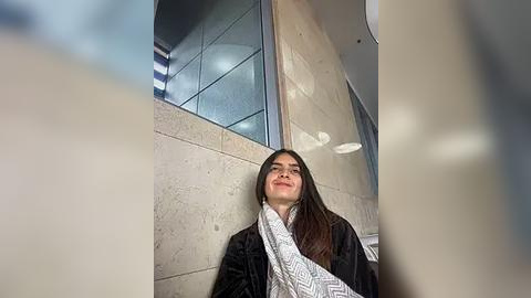 Media: Video of a young woman with long dark hair, wearing a black jacket and white patterned scarf, sitting against beige tiled wall in a modern, minimalist hallway.