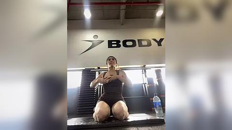 Media: A video of a person kneeling in a gym, wearing a grey sports bra and shorts, holding a phone for a selfie, with gym equipment and a water bottle in the background.