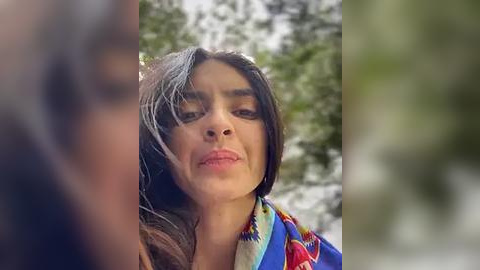 Media: Video of a young woman with long dark hair and a colorful, patterned shawl, captured in a forest with blurred greenery in the background.