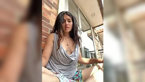 Media: Video of a distressed-looking woman with long dark hair, wearing a gray tank top and colorful shorts, sitting on a brick wall in broad daylight, blurred background.