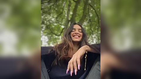 Media: A video of a smiling, light-skinned woman with long, wavy brown hair, wearing a black outfit, seated outdoors with blurred green foliage in the background.