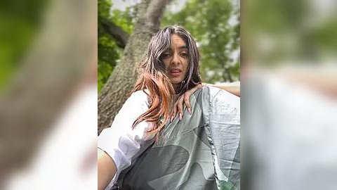 Media: Video of a wet woman with long, dark hair, wearing a white shirt and gray jacket, standing outdoors near a large tree.