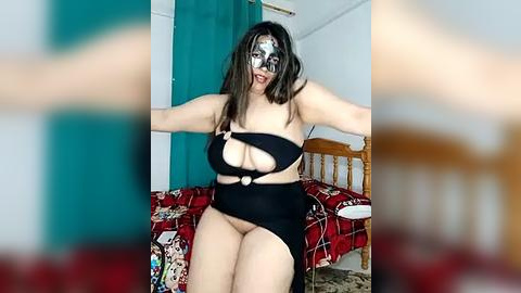 Media: Video of a Latina woman with long dark hair, wearing a black lace mask and revealing black lingerie, sitting on a bed with a red and white patterned blanket.