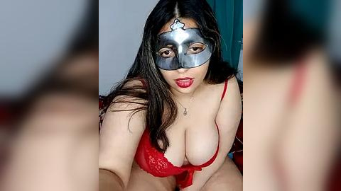 Media: Video of a curvy, fair-skinned woman with long dark hair, wearing a red lace bra and silver masquerade mask, sitting seductively.