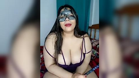 Media: A video of a Latina woman with long black hair, wearing a revealing purple lace bra, silver mask, and red lipstick, sitting on a bed with red and green floral sheets.